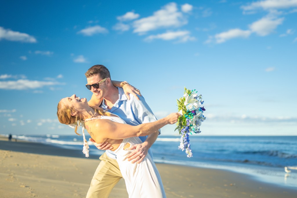 A newlywed couple shares a romantic kiss on the sandy shore of a wind-swept ocean, set against a brilliant blue sky dotted...