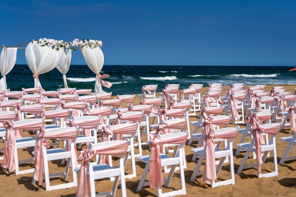 Beautiful pink and white wedding chairs set on a sunny day at the beach. A delicate white arch stands behind them, draped ...