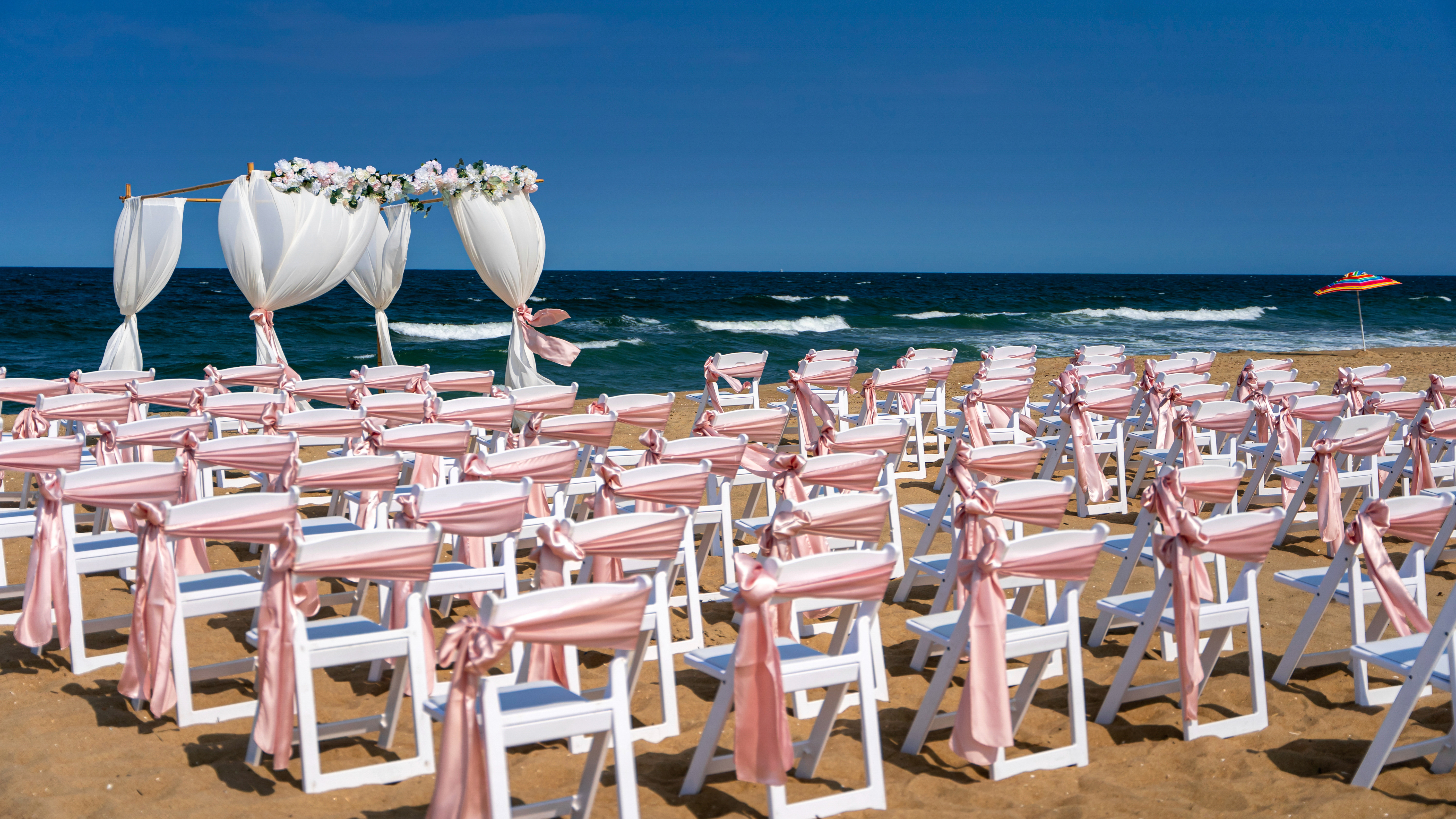 Beautiful pink and white wedding chairs set on a sunny day at the beach. A delicate white arch stands behind them, draped ...
