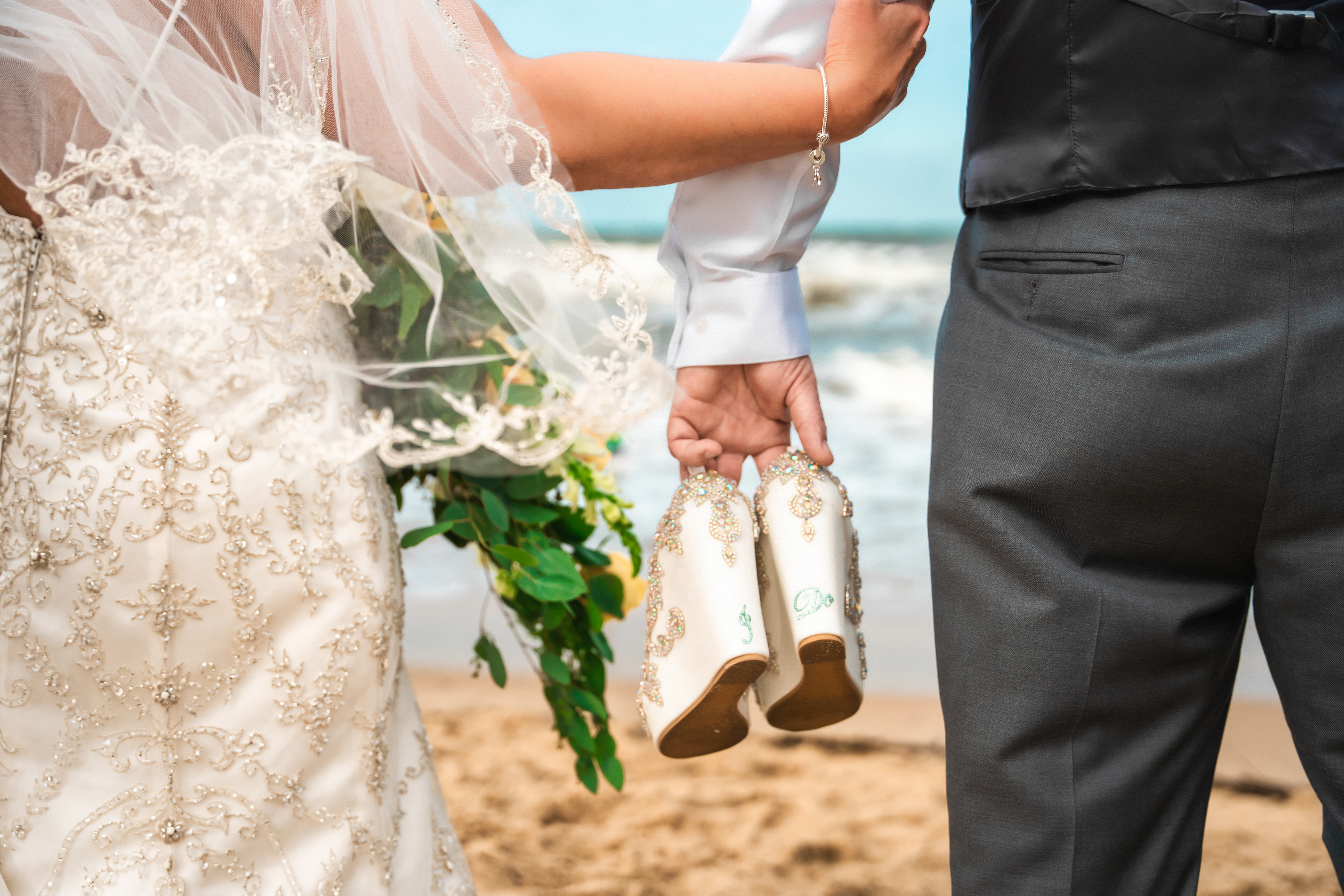 A newlywed couple standing on a sandy beach, with the ocean in the background. The bride is wearing a white dress with emb...